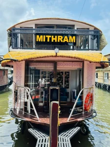 Straight front view of the MITHRAM01 premium 1-bedroom houseboat, showcasing its lower glass lounge and upper viewing deck floating on the Alleppey backwaters
