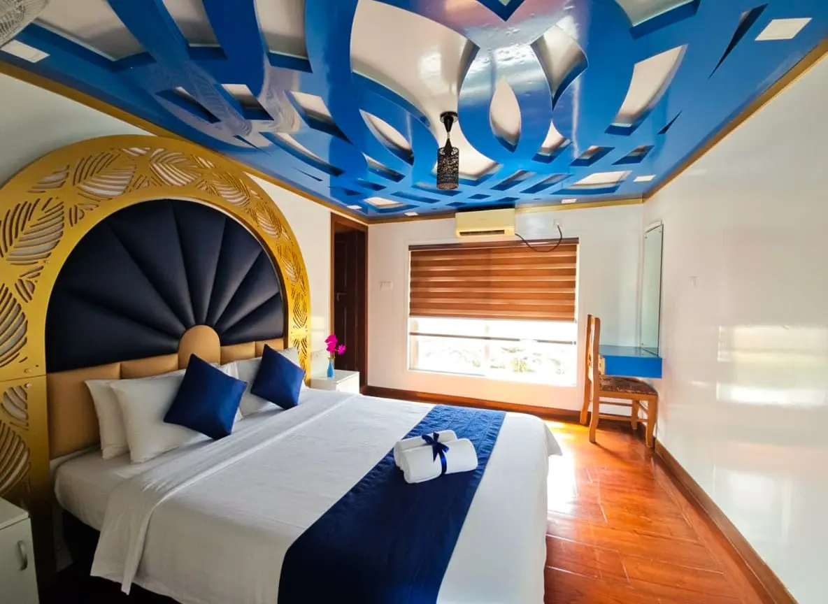 Royal Blue Interior Theme AC Bedroom – Houseboat Stay Front view of double bed with royal blue headboard, blue ceiling pattern, and side window with blinds.