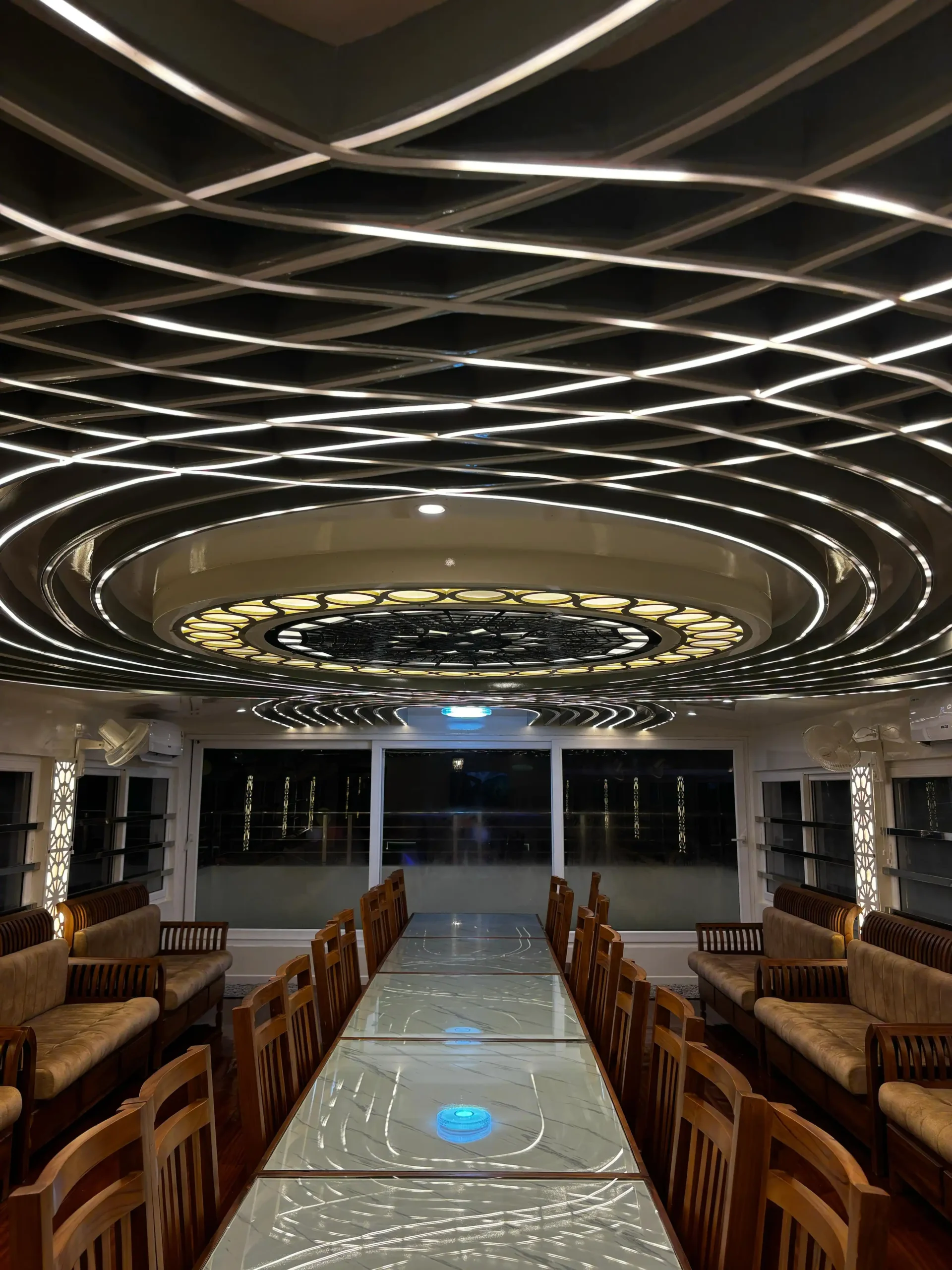 Nighttime Dinner Setup on Glass Upper Deck – Houseboat Stay Wooden dining table setup on the glass upper deck of Kingster 5 bedroom houseboat lit by glowing ceiling LED lights at night.